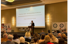 Prelegent stojący na scenie przy mikrofonie przedstawiający słuchaczom zagadnienie, w tle wyświetlana na ekranie prezentacja