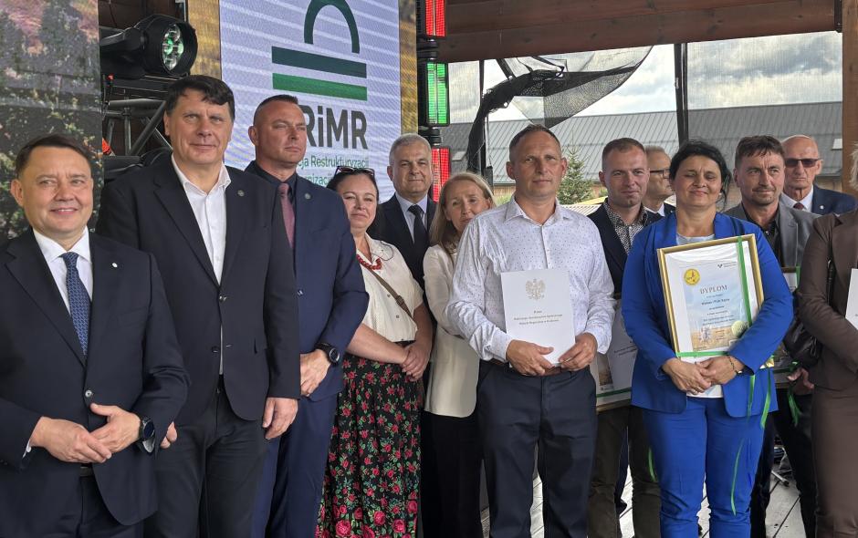 Na zdjęciu na scenie stoją mężczyźni i kobiety, sponsorzy i organizatorzy konkursu Bezpieczne Gospodarstwo Rolne. W pierwszym rzędzie kobieta i mężczyzna - zwycięzcy konkursu trzymają dyplomy
